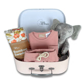 Baby gift set with pink outfit, bamboo swaddle, and plush elephant in a suitcase on a white background.